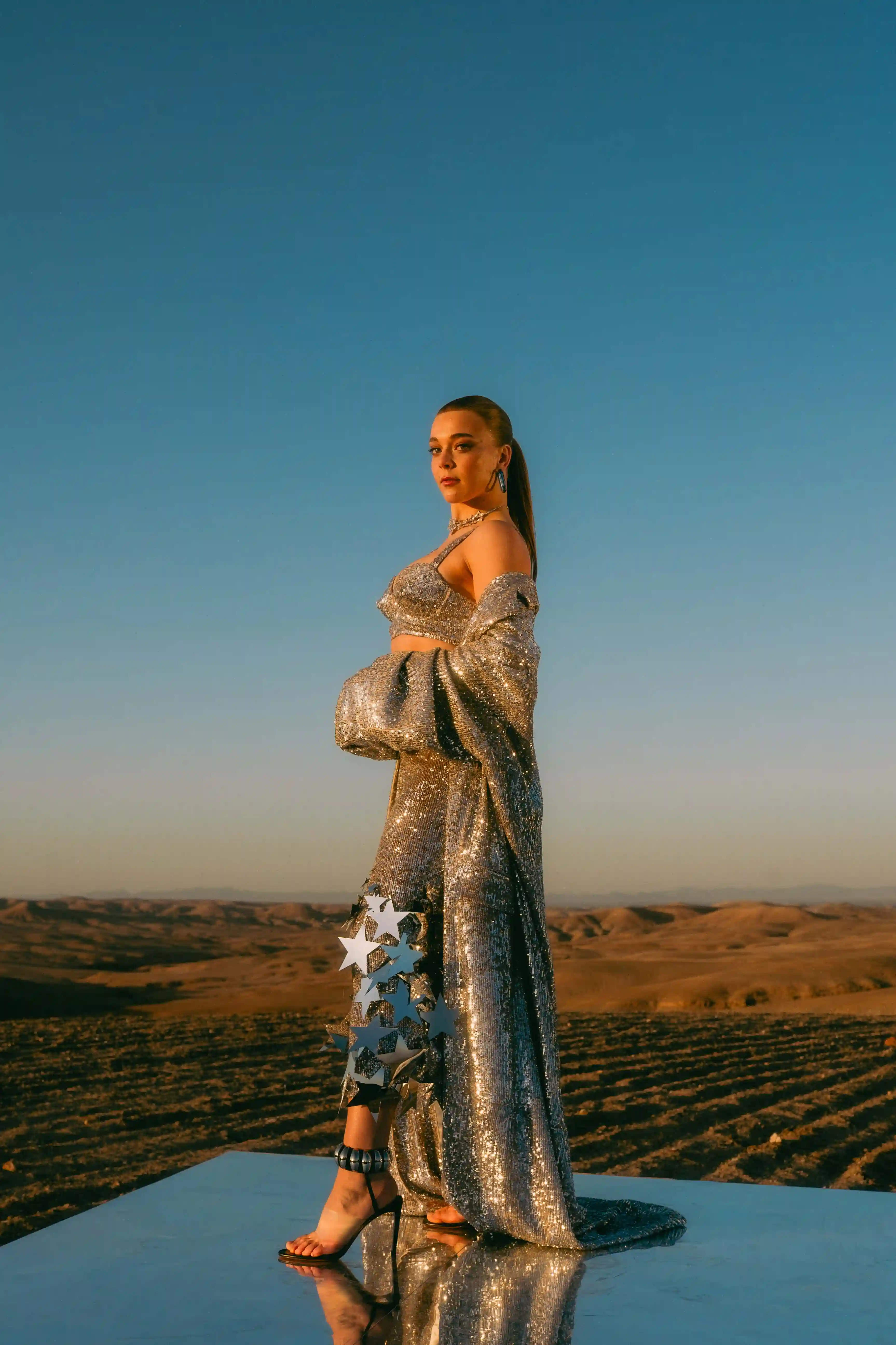 Becky Hill in a long dress ona mirrored platform in the desert